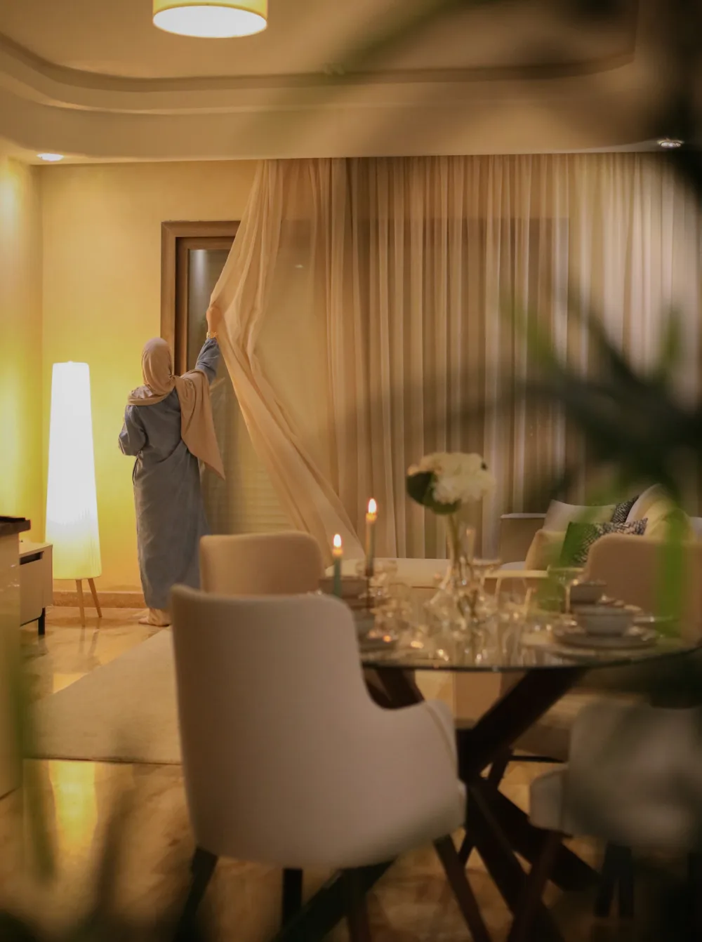 Myriam Monfort, de dos, ajuste un rideau dans un salon qu'elle a aménagé, table dressée au premier plan.