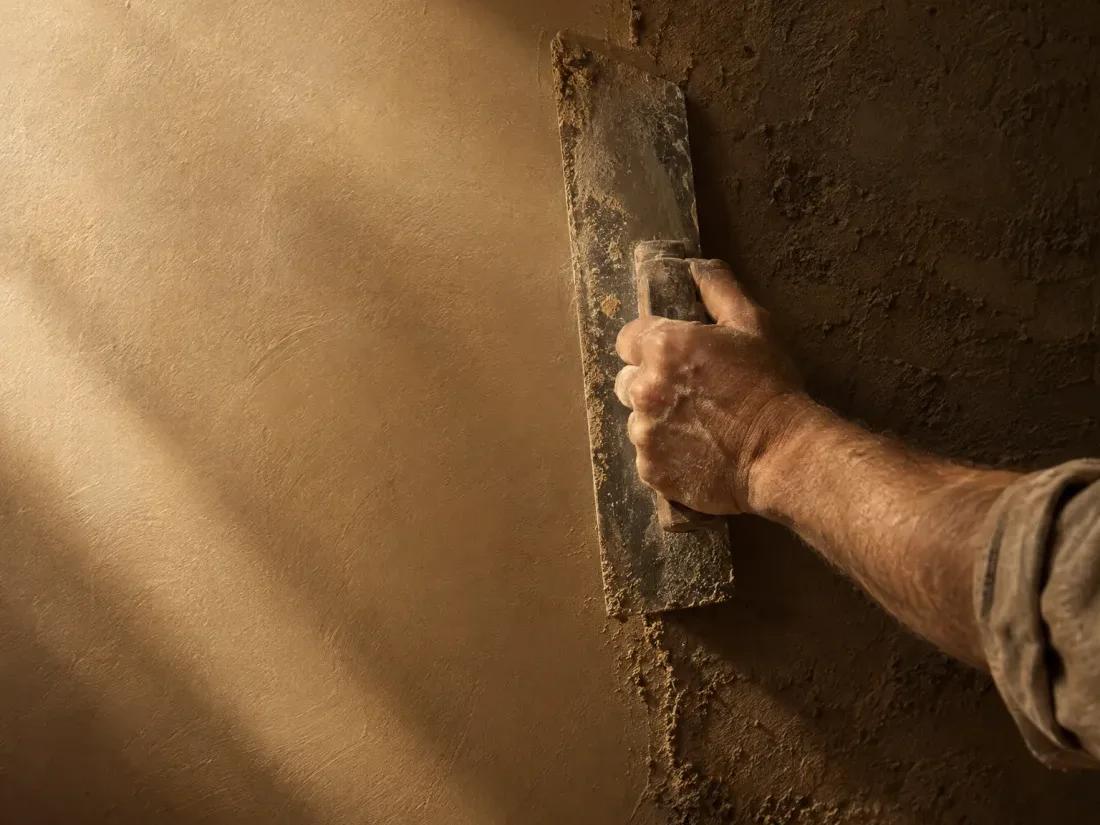 La main d'un artisan qui lisse un enduit beige à la taloche, dans une lumière rasante.